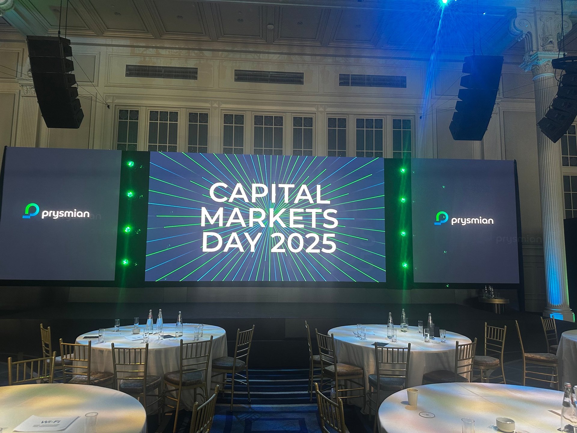 Event venue setup for Capital Markets Day 2025 with large display screens, round dining tables, and stage lighting