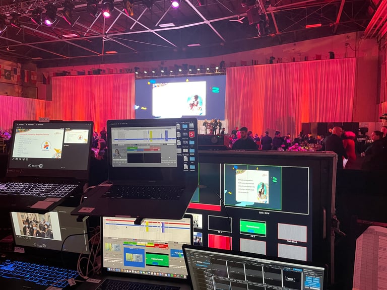 Professional event production setup with multiple computer monitors, red stage lighting, projection screen and audience silhouettes